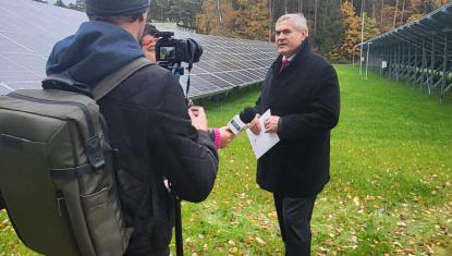 Ekipa telewizyjna nagrywa rozmowę z mężczyzną w czarnym płaszczu, w tle panele fotowoltaiczne i las w jesiennej szacie