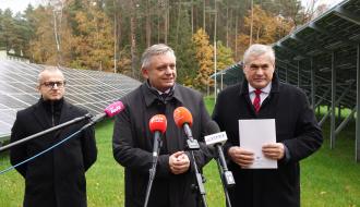 Trzech mężczyzn stoi przed mikrofonami na statywach, w tle panele fotowoltaiczne i jesienny las