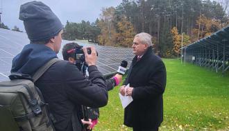 mężczyzna w ciemnym ubraniu udziela wywiady do kamery, w tle panele fotowoltaiczne i jesienny las